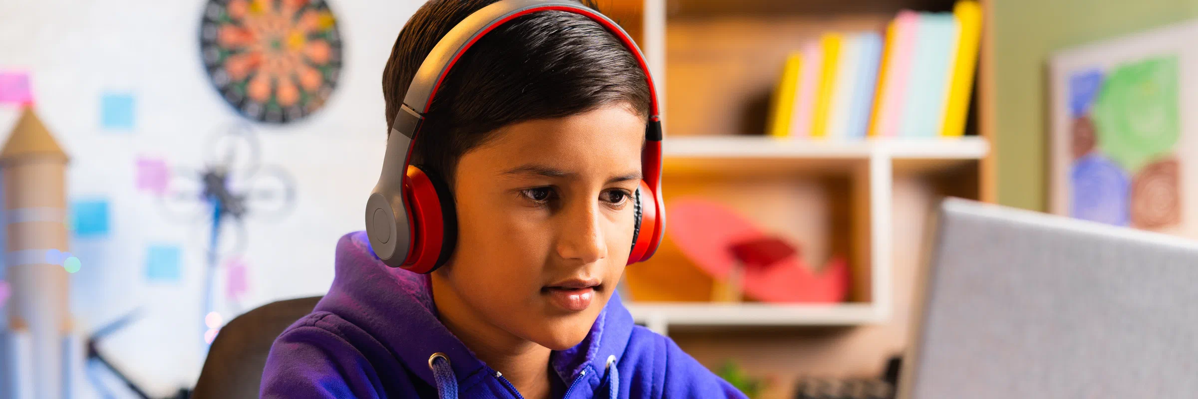 A school-age boy in headphones working on a laptop at a desk in his bedroom