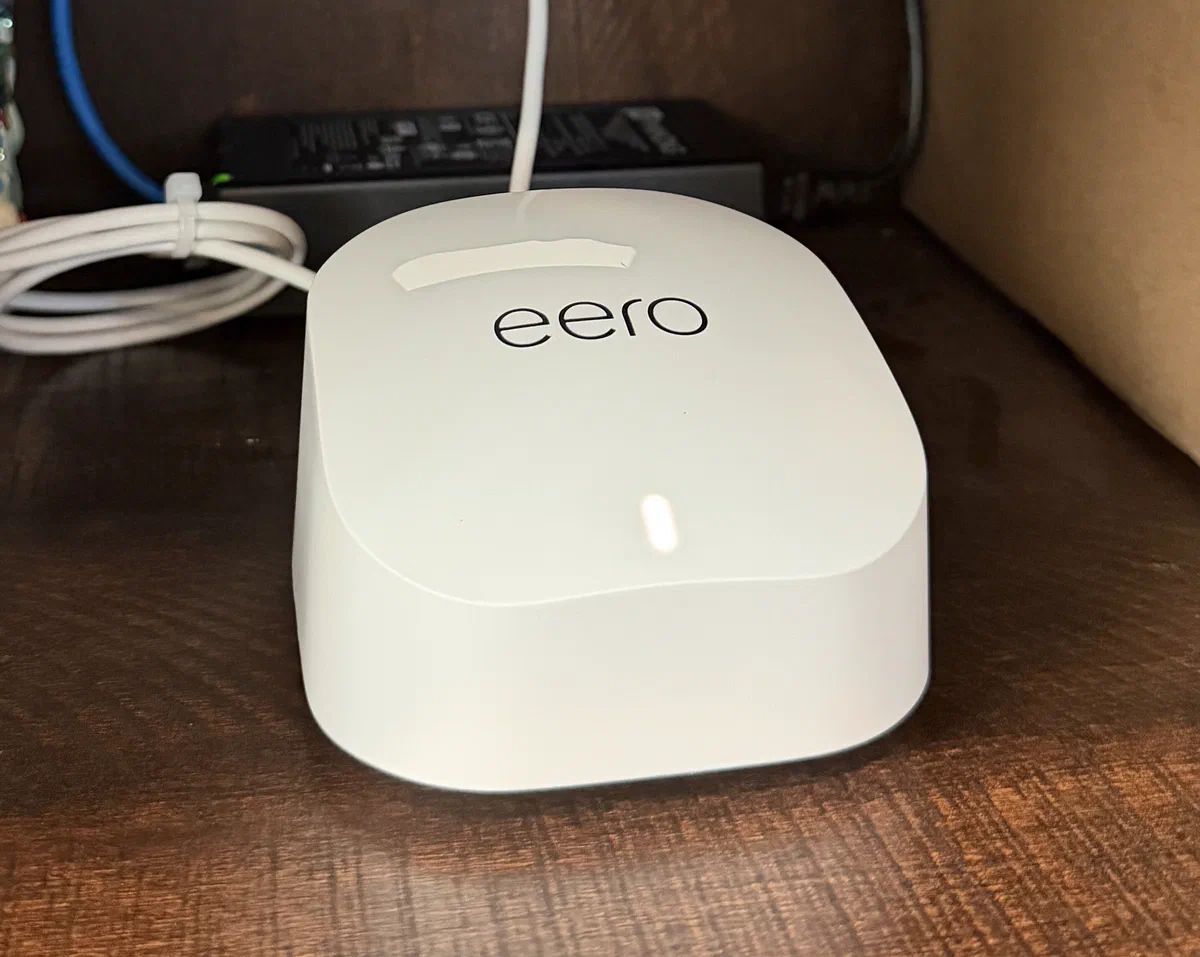 A white eero mesh Wi-Fi unit sitting on a wood shelf with its status light lit and a network cable coiled behind it