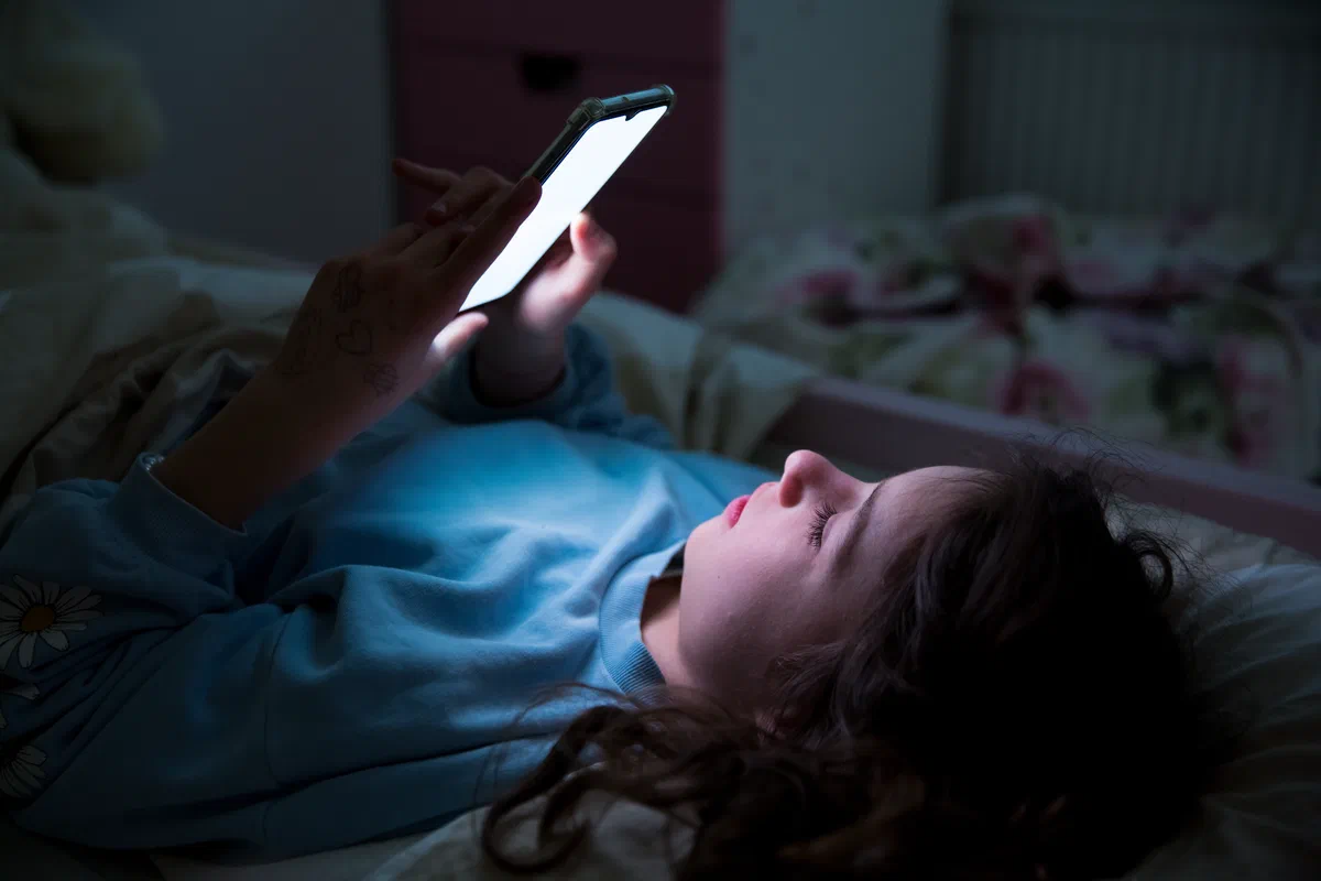 A young girl lying in bed in a dark room, her face lit by the screen of the phone she is holding above her