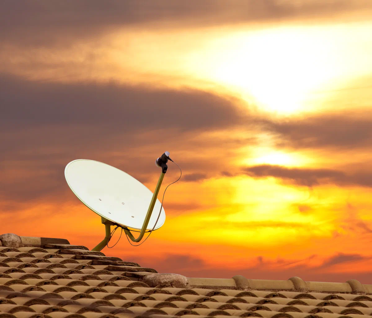 An ageing satellite internet dish still mounted on a Florida tile roof, silhouetted against an orange sunset