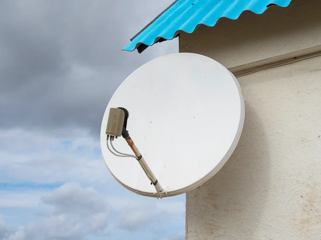 An old white satellite dish bolted to the stucco wall of a house under a blue metal roof edge, its mount arm rusting and cables zip-tied down the arm